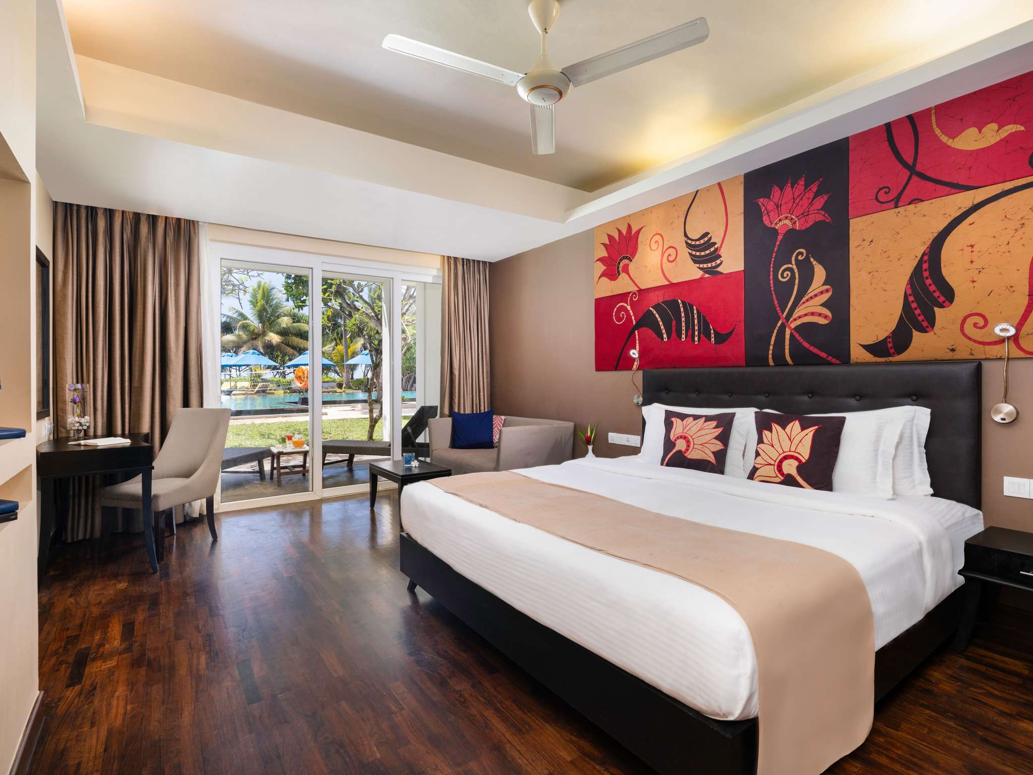 A Superior Garden View King Bed room: a king bed under a red, gold and black painted panel, a sofa, a desk and a dark wooden floor, and glass doors open to a patio with loungers, a lawn and the pool