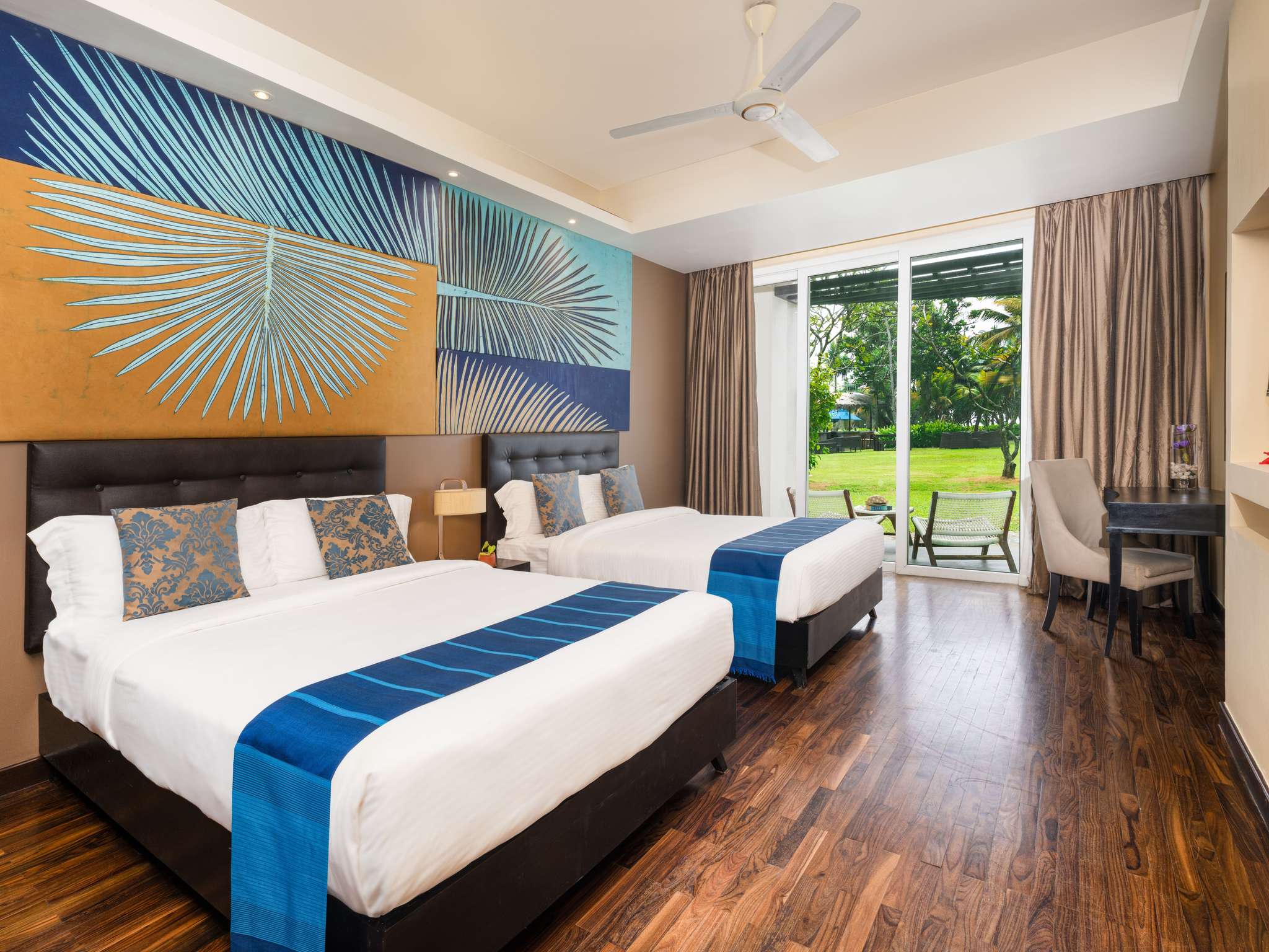 A Superior Garden View Twin Bed room: 2 beds with blue runners under a painted palm-leaf panel, a desk, and glass doors open to a patio with 2 chairs and a lawn under palms