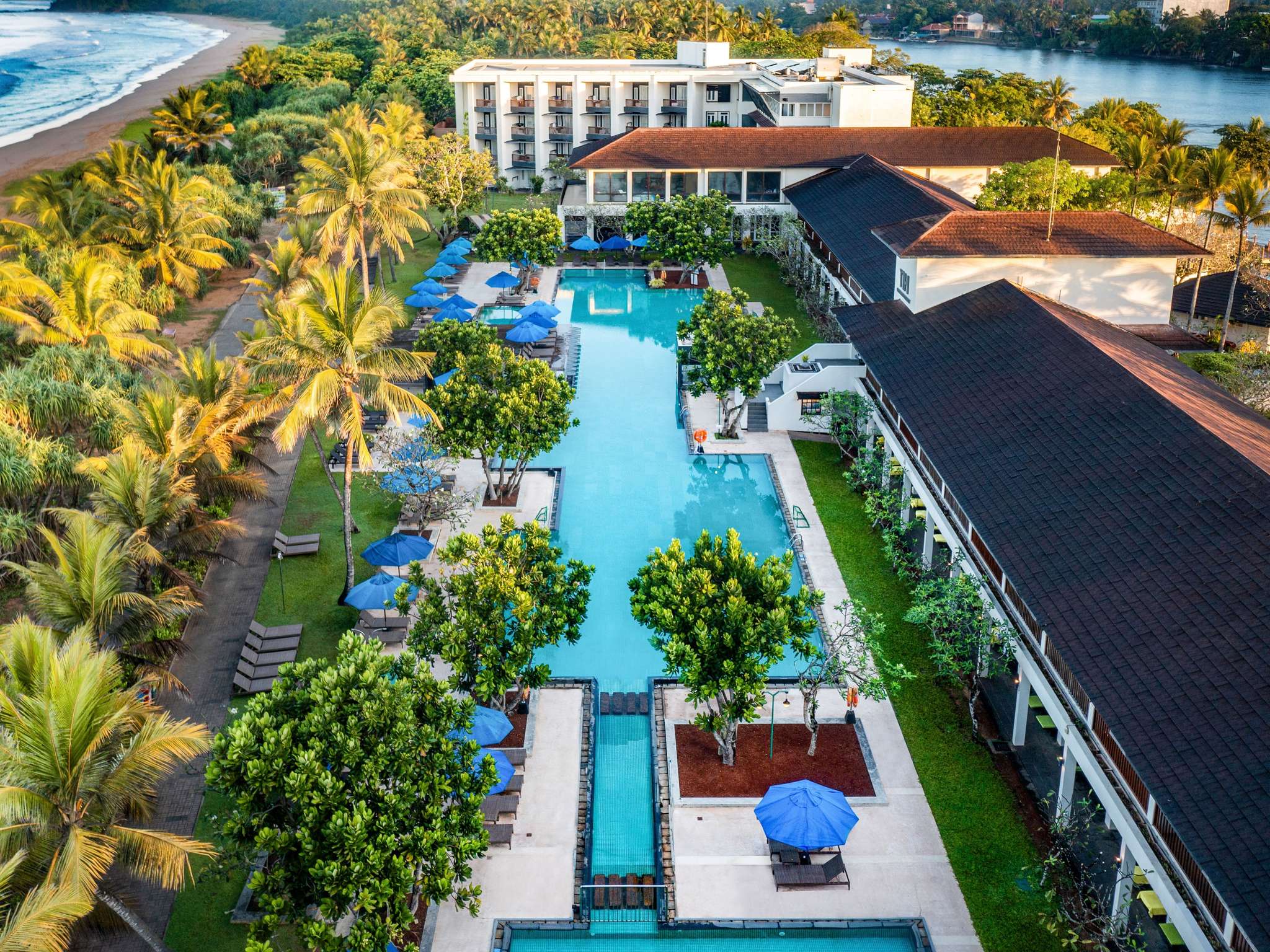 The resort from the air: a long pool running between 2 tiled-roof wings and lawns, palms along the beach on the left and a white block of rooms with the river behind it