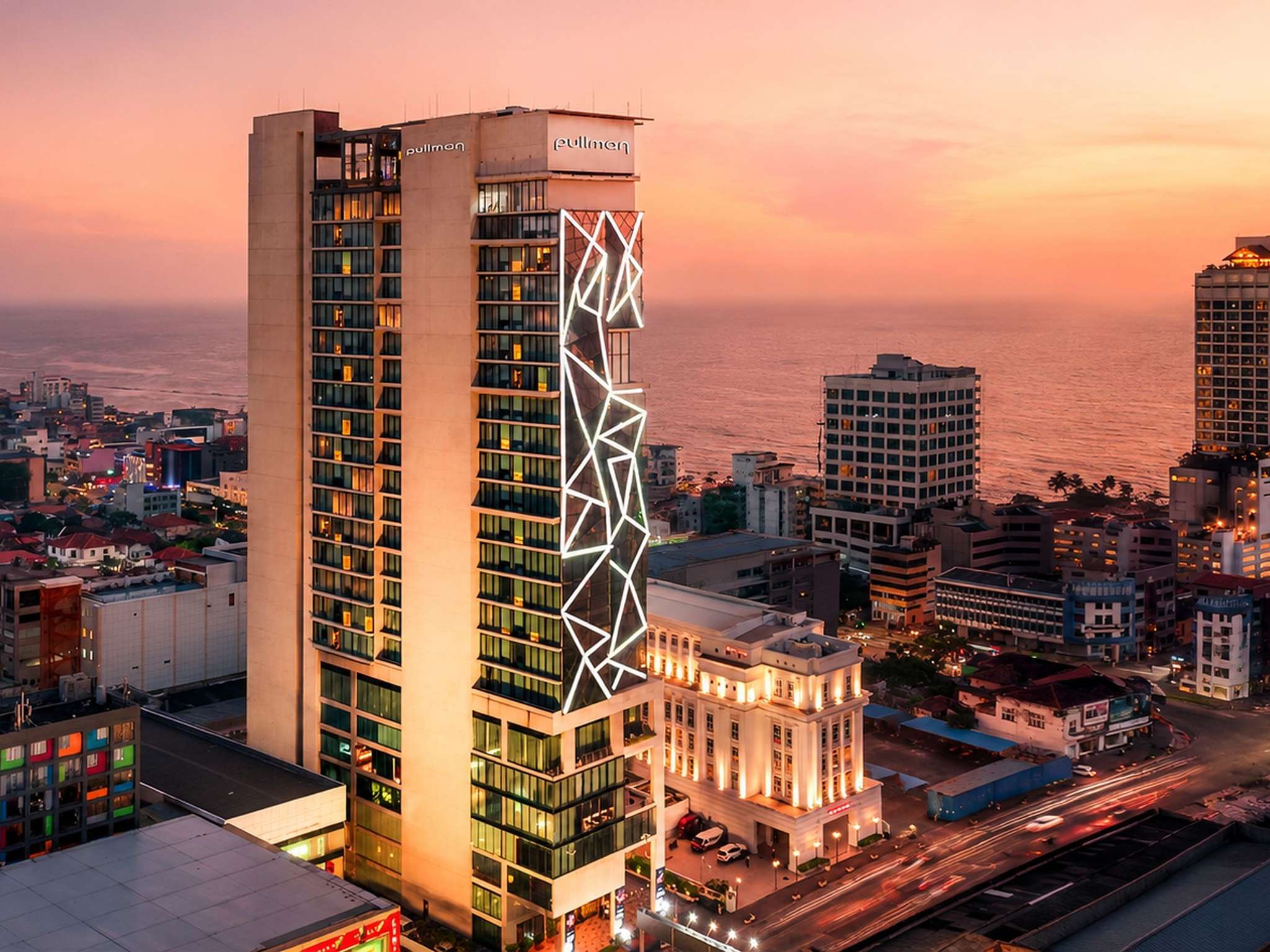 The hotel tower at dusk with a lit geometric pattern on its glass, a white colonnaded building at its foot, and the city and the sea behind