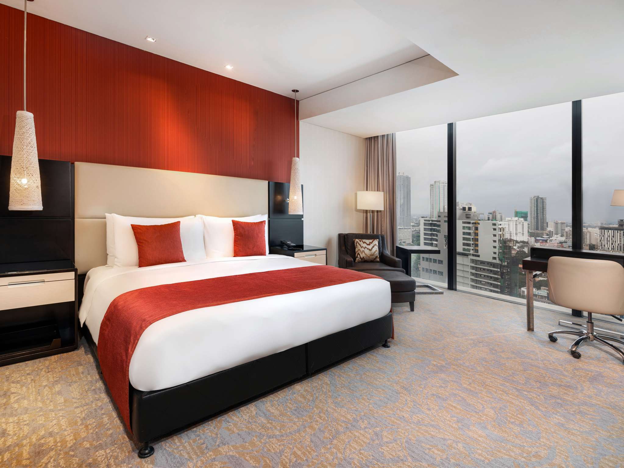 A Superior City View room: a king bed with a red throw against a red feature wall, woven pendant lamps, a leather armchair, a desk, and floor-to-ceiling windows over city towers